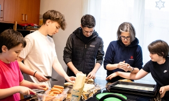 Mutter und vier Kinder zwischen Grundschulalter und Teenageralter stehen gemeinsam am Küchentisch und bereiten Essen zu