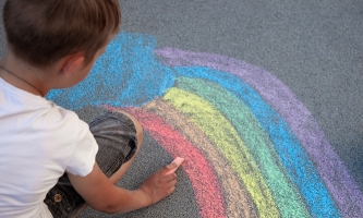 Ein Junge in weißem Shirt malt mit Straßenkreide einen Regenbogen auf den Asphalt.