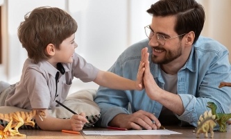 Vater und Sohn liegen lachend mit Papier, Stiften und Dino-Figuren auf dem Boden und machen High five