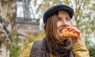 Frau mit Baskenmütze und halblangen dunkelblonden Haaren beißt in Croissant vor Eiffelturm