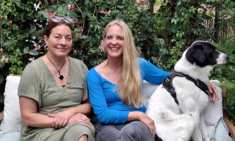 Julia Dahmen und Caro Matzko lächeln in die Kamera. Caro Matzko hat ihren Arm dabei um einen schwarz-weißen Hund gelegt.