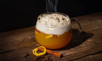 Dampfender Punsch in gläserner Tasse mit Sahne auf Holztisch, daneben Zimtstange und Zitronenschalenring