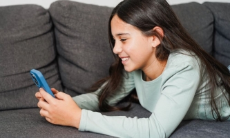 Kind mit langen Haaren liegt fröhlich auf Sofa und schaut in Handy