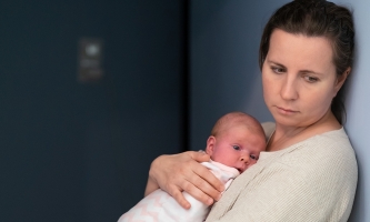 Eine Frau blickt nachdenklich auf den Boden. Dabei hält sie ein Baby auf dem Arm.