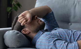 Mann liegt krank auf Sofa und hat den Arm über das Gesicht gelegt