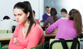 Eine Jugendliche mit langen braunen Haaren und einem eine hellroten Longsleeve blickt traurig und angespannt in die Kamera. Sie kehrt ihrer Klasse den Rücken zu, die im Hintergrund den Unterricht verfolgt.