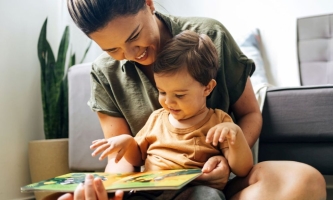 Mutter und kleines Kind sitzen fröhlich vor der Couch auf dem Boden und schauen zusammen ein Bilderbuch an