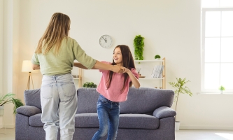 Eine Mutter tanzt in einem Wohnzimmer vor dem Sofa mit ihrer etwa zwölfjährigen Tochter.