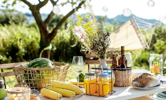 Sommerlich gedeckter Tisch im Freien mit Obst, Getränken, Brot und Blumen, im Hintergrund Lampions und eine Hängematte