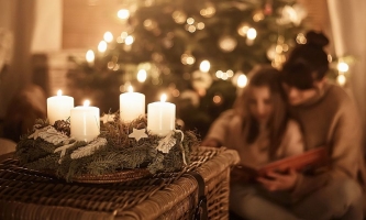 Adventskranz mit vier brennenden Kerzen und leuchtender Weihnachtsbaum im Wohnzimmer, Mutter und Kind schauen ein Buch an