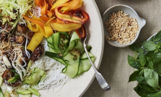 Salat mit Karotten, Gurken, Fleisch und Reisnudeln auf Teller, daneben Schälchen mit Thai-Basilikum und Erdnüssen