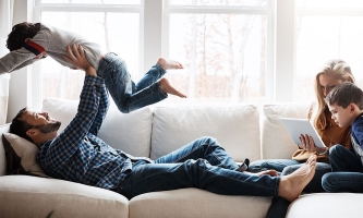 Vater auf Sofa lässt Kind über sich fliegen, Mutter schaut mit anderem Kind auf Laptop