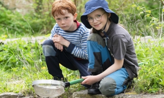 zwei Kinder in der Natur mit Sieb knien im Gras und lachen in die Kamera