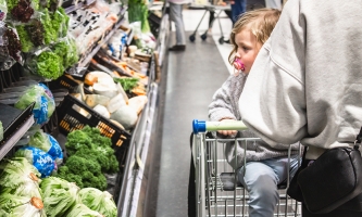 Mutter mit Kind und leerem Einkaufswagen vor einem Regal mit Gemüse im Supermarkt