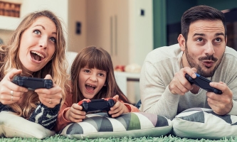 Mutter, Vater und Kind liegen mit Spielkonsolen in den Händen auf dem Teppich und schauen begeistert