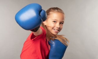 Ein Mädchen kickt mit ihren blauen Boxhandschuhen und lächelt dabei in die Kamera.