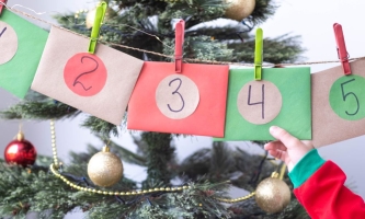 Kinderhand hält Umschläge eines hängenden Adventskalenders, im Hintergrund ein Christbaum 