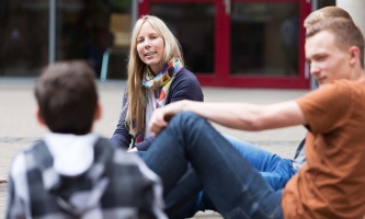 Eine junge Frau sitzt auf dem Boden eines Pausenhofs umringt von Jugendlichen und hört aufmerksam zu.