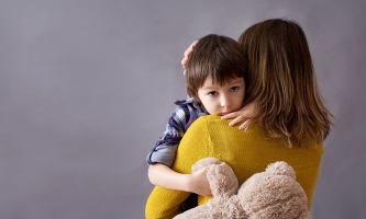 Eine Mutter hält ihren Sohn auf dem Arm. Sie ist dem Bild abgewandt, er hält seinen Teddy fest und schaut in die Kamera.