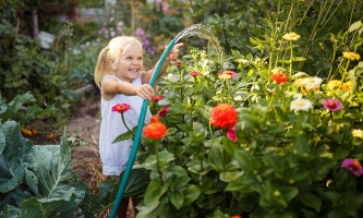 Ein kleines Mädchen steht mitten im Garten, lacht und gießt mit dem Gartenschlauch die Blumen.