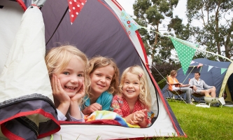 Drei kleine Mädchen liegen im Zelt und schauen lächelnd heraus. Im Hintergrund sieht man die Eltern auf Klappstühlen sitzen.