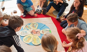 Kinder sitzen in Kreis auf Boden und beugen sich über Blüte aus Papierbildern