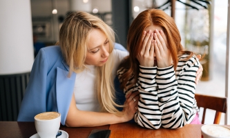 Zwei Frauen sitzen in einem Café. Eine Frau hält sich die Hände vor die Augen, die andere nimmt sie in den Arm.