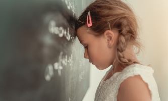 Ein Mädchen steht mit traurigem Blick vor einer Schultafel und lehnt seine Stirn daran.