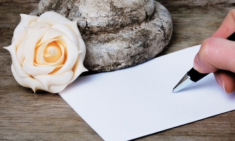 Hand mit Stift schreibt Trauerkarte, daneben Stein und cremeweiße Rose 