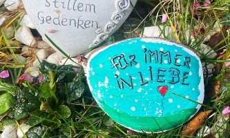 Mit bunten Farben bemalter Stein mit Schrift "Für immer in Liebe" auf einem Grab 