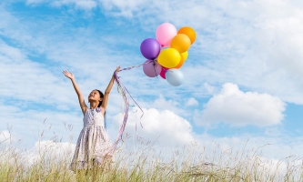 Kind in Sommerkleid auf Wiese hält Luftballons hoch in die Luft und streckt die Hände zum Himmel