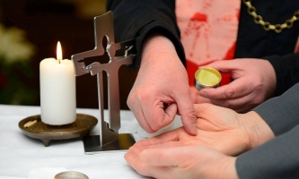 Ein Priester salbt einen Kranken mit Krankenöl, daneben ein Kreuz und eine brennende Kerze 