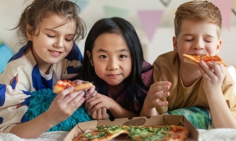 Drei Kinder liegen zusammen auf Bett und essen Pizza aus Karton 