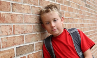 Junge mit Schulrucksack lehnt schüchtern an Mauer 