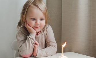 Mädchen schaut am Namenstag glücklich auf brennende Kerze vor ihm auf dem Tisch 
