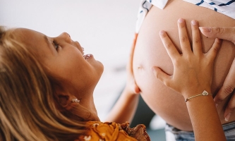 Kind legt Hände auf Bauch einer schwangeren Frau und schaut fragend und glücklich nach oben 