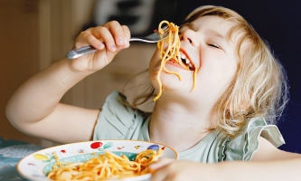 Kind lässt Spaghetti mit Soße von der Gabel in den Mund hängen 