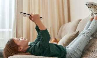 Kind liegt mit den Füßen nach oben auf dem Sofa und schaut auf ein Tablet