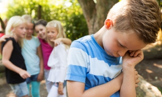 Junge traurig auf dem Schulhof, tuschelnde Gruppe Mädchen im Hintergrund