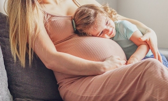 Schwangere Frau auf Sofa, Kleinkind legt Kopf auf ihren Bauch