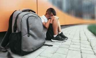 Junge mit Rucksack sitzt mit vor das Gesicht geschlagenen Händen auf dem Boden vor dem Schulgebäude 