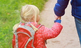 Kindergartenkind mit Rucksack geht an Hand des Vaters einen Weg entlang 