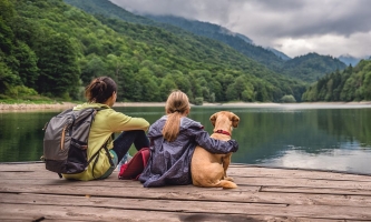 Frau, Kind und Hund sitzen auf Holzplanken und schauen auf einen Bergsee 