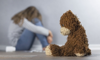 Ein Teddy sitzt im Vordergrund mit hängendem Kopf, hinter ihm - verschwommen zu sehen - sitzt ein in sich zusammengekauertes trauriges Mädchen.