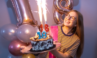 Ein Mädchen hält eine Torte in der Hand. Auf der Torte sind Geburtstagskerzen in Form einer 18, sprühendes Feuerwerk – und im Hintergrund sind Luftballons als Zahl 18 zu sehen.