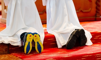 Es sind die Beine zweier knieender Ministranten zu sehen. Unter ihren Gewändern schauen die Schuhe heraus.