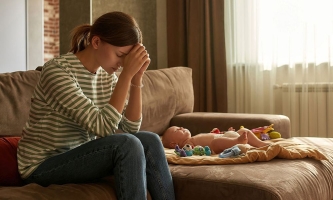 Mutter auf dem Sofa neben Säugling stützt bedrückt den Kopf auf ihre Hände 