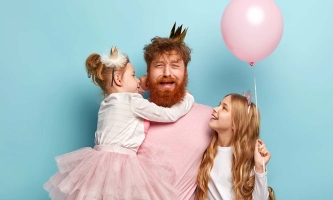 Ein Papa im rosa T-Shirt und mit goldener Krone hält seine eine Tochter im rosa Tütü auf dem Armm die andere Tochter steht mit einem rosa Luftballon neben ihm. Der Papa macht ein verzweifeltes Gesicht.