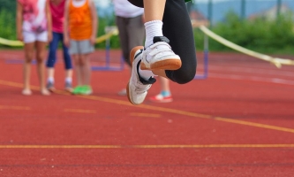 Kinderfüße in der Luft beim Weitsprung auf dem Sportplaz, im Hintergrund wartende Kinder