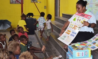 Lehrerin zeigt auf dem Boden sitzenden Kindern eine Bildtafel 